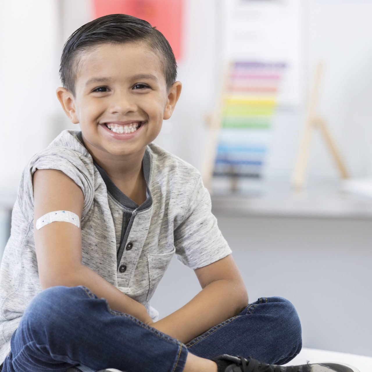 An adorable little elementary age boy sits cross legged on an exam table in his pediatrician's office. He displays an arm bandage as he smiles for the camera.